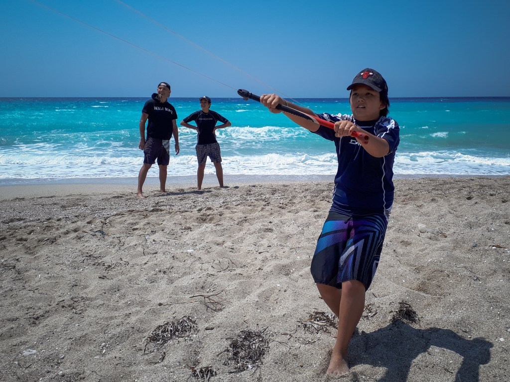 A young boy learning to kitesurf on the beach with people in the back and clear blue and wavy sea