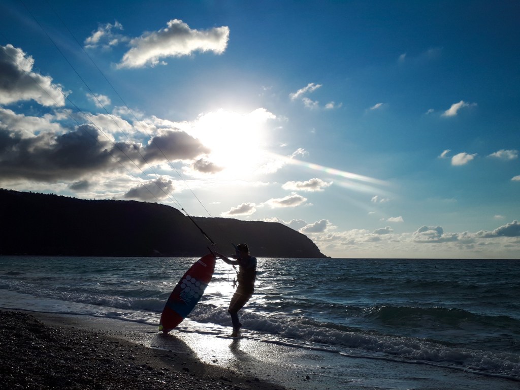 A kitesurfer on the Agios Ioannis beach in Lefkada with his harness and kitebar in one hand, surfboard in the other hand, ready to go kitesurf in the sea